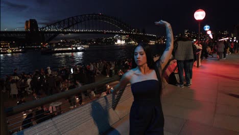 Mujer-Bailando-Con-Vestido-Negro---Mujer-Joven-De-Piel-Oscura-Con-Cabello-Largo-Y-Vestido-Negro-Bailando-Gratis-En-El-Recinto-De-La-ópera-De-Sydney-Por-La-Noche,-Como-Telón-De-Fondo-Del-Puente-Del-Puerto-De-Sydney---4k,-59