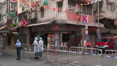 Trabajadores-De-La-Salud-Vestidos-Con-Trajes-Para-Orinar-Y-Un-Oficial-De-Policía-Montan-Guardia-Frente-A-Una-Barrera-En-Un-área-Bloqueada-Para-Contener-La-Propagación-Del-Brote-De-La-Variante-Del-Coronavirus-En-Hong-Kong