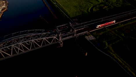 Panorámica-Lateral-Aérea-Que-Muestra-Un-Puente-Levadizo-De-Acero-Sobre-El-Río-Ijssel-Con-Un-Tren-Que-Pasa-Al-Atardecer