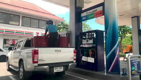 Chico-De-Gasolina-Cargando-Petróleo-En-Bidones-De-Metal-Rojo-En-La-Parte-Trasera-De-Un-Coche-Isuzu-Dmax-En-La-Gasolinera-En-Bangkok,-Tailandia---Tiro-Completo