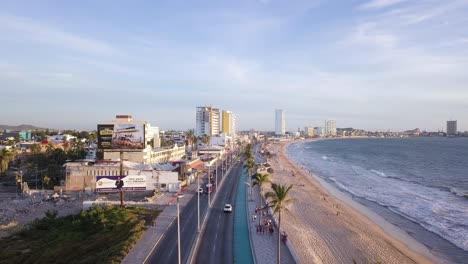 Volando-Sobre-El-Paseo-Marítimo-De-La-Playa-Del-Malecón-En-Mazatlán-México-Antes-Del-Atardecer,-El-Tráfico-De-Automóviles-Y-La-Gente-Caminando-Por-El-Paseo-Marítimo-Costero-Junto-A-La-Playa-De-Arena-Y-El-Mar,-El-Paisaje-Urbano-Costero