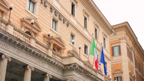 Tiro-De-ángulo-Bajo-Del-Edificio-Administrativo-De-Piazza-Colonna-A-Lo-Largo-De-La-Bandera-Italiana-Y-Del-Euro-En-Roma,-Italia-Durante-El-Día