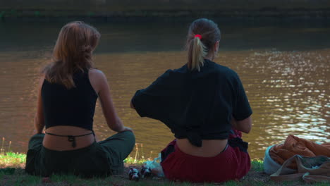 Young-girls-enjoying-summer-afternoon-on-riverside,-while-boys-passing-by-on-a-small-boat