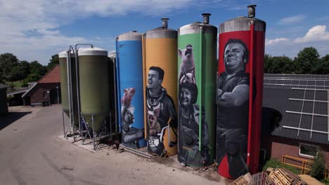 Aerial-of-Dutch-graffiti-decoration-drawing-of-farmers-family-with-animals-on-large-farm-stock-silo-containers-contrasted-against-a-blue-sky-with-fluffy-clouds