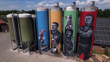 Closeup-aerial-pan-around-graffiti-decoration-drawing-of-farmers-family-with-animals-on-large-farm-stock-silo-containers-contrasted-against-a-blue-sky-with-fluffy-clouds