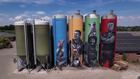 Ascending-aerial-movement-of-graffiti-decoration-drawing-of-farmers-family-with-animals-on-large-farm-stock-silo-containers-contrasted-against-a-blue-sky-to-top-down