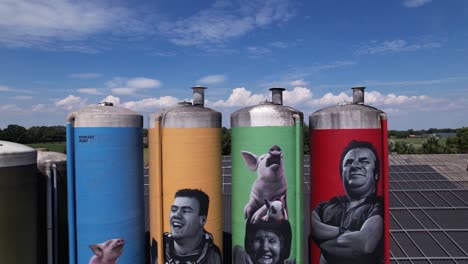 Antena-De-Colorido-Dibujo-De-Decoración-De-Graffiti-De-La-Familia-De-Agricultores-Con-Animales-En-Grandes-Contenedores-De-Silo-De-Ganado-Contrastado-Con-Un-Cielo-Azul-Con-Nubes-Esponjosas