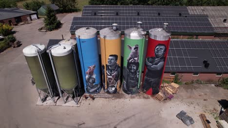 Aerial-descend-reveal-graffiti-decoration-drawing-of-farmers-family-with-animals-on-large-farm-stock-silo-containers-contrasted-against-a-blue-sky-with-fluffy-clouds