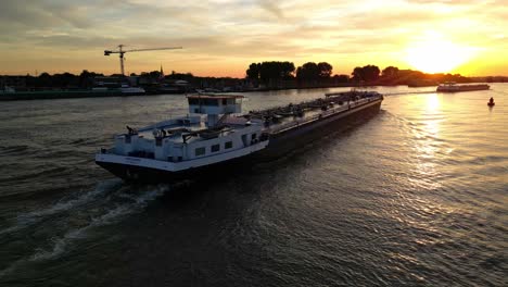 Aerial-View-Of-Maxima-Inland-Liquid-Tanker-Navigating-Along-Oude-Maas-During-Bright-Orange-Yellow-Sunset-Skies