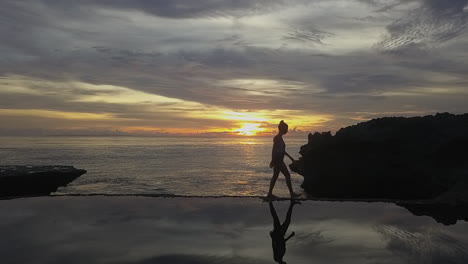 Mujer-Slo-Mo-Reflejada-En-Una-Piscina-Natural,-Camina-A-Través-De-La-Puesta-De-Sol-Del-Océano