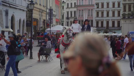 En-Una-Calle-De-Cracovia,-Polonia,-Donde-Mucha-Gente-Camina,-Dos-Mujeres-Conducen-Un-Carruaje-Tirado-Por-Caballos-Tirado-Por-Dos-Caballos-Blancos