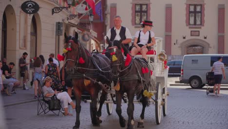 Two-men-driving-a-horse-carriage-which-is-pulled-by-two-horses-in-the-street-of-Kraków,-Poland-where-numerous-people-are-walking-in-the-street