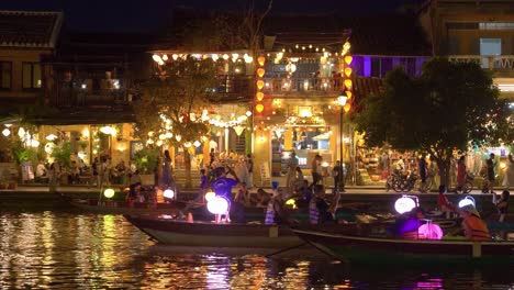 Paseos-Nocturnos-En-Barco-Para-Turistas-En-El-Río-Hoai-En-La-Antigua-Ciudad-De-Hoi-An,-Faroles-Que-Iluminan-Edificios-Coloniales-Históricos-Que-Bordean-El-Paseo-Marítimo