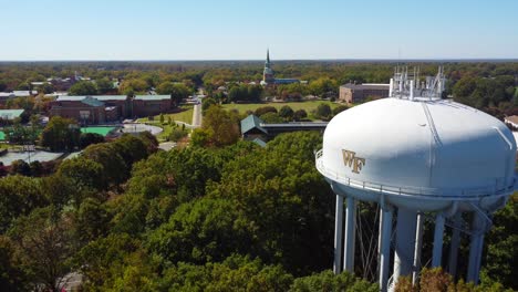 Antena-Hacia-Atrás-En-La-Torre-De-Agua-En-La-Universidad-Wake-Forrest-Con-Capilla-De-Espera-En-El-Fondo,-Winston-salem,-Nc