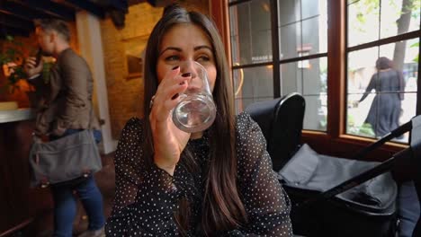 Long-haired-female-drinking-beverage-in-trendy-London-bar-sitting-next-to-the-window-with-people-passing-in-the-background