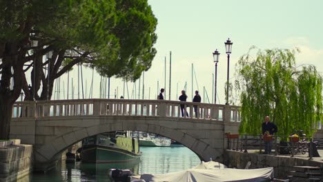 Mujer-Cruzando-Un-Viejo-Puente-De-Piedra-Cerca-De-Un-Pequeño-Puerto-De-Un-Lago