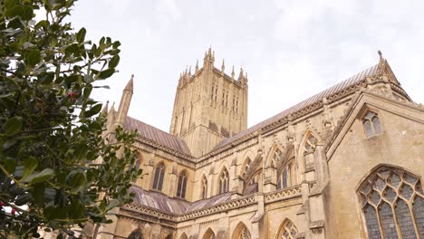 Outside-walls-and-tower-of-Wells-Cathedral,-camera-rotating-to-the-right-from-behind-a-tree-revealing-the-rest-go-the-Cathedral