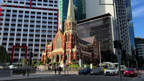 Static-shot-at-downtown-Brisbane-city,-pedestrians-crossing-the-street,-cars-driving-pass-heritage-listed-Victorian-Gothic-Revival-architecture,-Albert-street-uniting-church-in-the-background