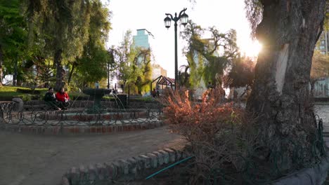 Couple-sitting-on-a-bench-in-a-small-square-with-fountain-in-a-city-garden-at-sunset