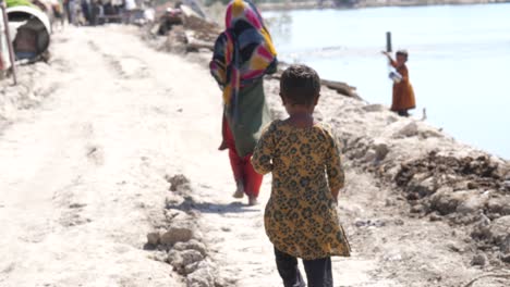 Video-De-Una-Niña-Pequeña-Corriendo-Descalza-En-El-Camino-Roto-Para-Seguir-A-Su-Madre-Mientras-Camina-Frente-A-Su-Maher,-Sindh