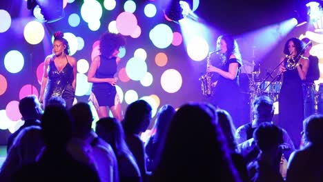 High-on-Heels-ladies-Group-play-on-stage-with-colorful-background-animation-with-people-enjoying-the-music-in-front-of-them,-static-slow-motion-view