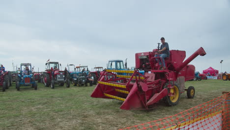 Cosechadora-Massey-Ferguson-Roja-Conduciendo-En-El-Campo-Durante-La-Exposición-Con-Tractores-Agrícolas-Clásicos-Alineados-En-Segundo-Plano.