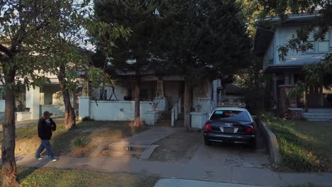 Aerial-drone-shot-over-a-police-car-parked-in-an-open-garage-outside-a-house-with-passerby-walking-along-the-sidewalk-during-evening-time