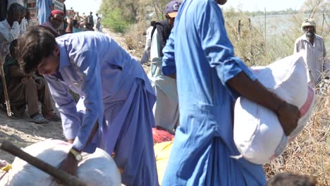 Locals-Helping-To-Carry-Sacks-Of-Aid-Beside-River-At-Flood-Relief-Drive-In-Maher,-Sindh