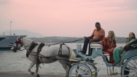 Eine-Hellblaue-Traditionelle-Kutsche-Mit-Einem-Weißen-Pferd-Und-Touristen,-Die-Vor-Dem-Grand-Poseidonion-Hotel-Bei-Einem-Romantischen-Roten-Sonnenuntergang-Am-Abend-Des-Küstenhafenpiers-Von-Spetses-Vorbeifahren