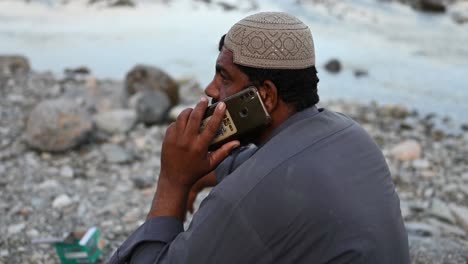 Hombre-Musulmán-Paquistaní-Se-Sentó-Al-Lado-Del-Río-Hablando-Por-Teléfono-Móvil-En-Sindh