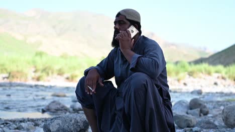 Hombre-Musulmán-Paquistaní-Se-Sentó-Al-Lado-Del-Río-Hablando-Por-Teléfono-Móvil-Con-Un-Cigarrillo-En-La-Mano-En-Sindh