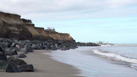 Two-people-walk-by-sea-defences-of-granite-rocks-defending-the-Norfolk-coastline-damaged-by-coastal-erosion-on-a-beach-as-waves-roll-in-from-the-North-Sea