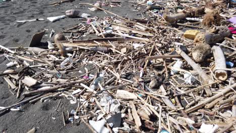 Piles-of-Trash-and-Wood-on-Black-Sand-Bali-Beach-near-Biaung-Denpasar,-Indonesia-flooded-rivers-brings-plastic-rubbish-to-the-Ocean,-60-fps