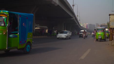 El-Tráfico-Se-Cierra-En-La-Carretera-Bajo-El-Sobrevuelo,-Pasando-Motos-Y-Coches,-Bicicletas,-Autobuses,-Rickshaw,-Vallas-Publicitarias-Y-Edificios-En-El-Fondo