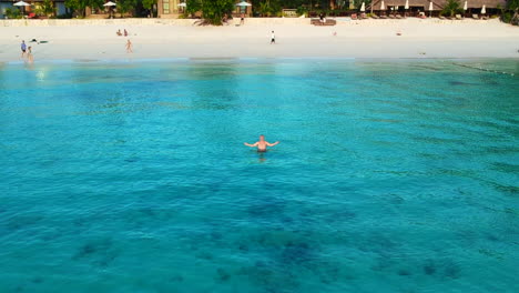 An-aerial-shot-of-a-man-standing-into-the-water-of-an-ocean-with-few-people-in-the-background