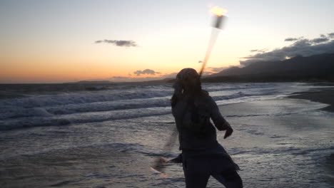 A-male-flow-art-dancer-in-silhouette-performing-his-fire-spinning-on-the-beach-at-sunset-with-ocean-waves-in-slow-motion