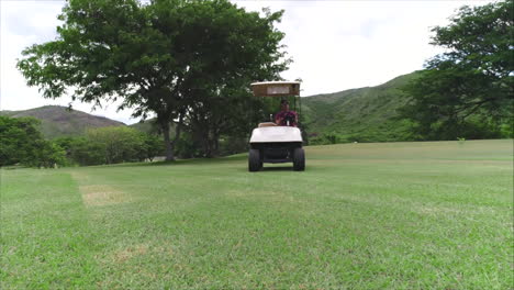 Cámara-Lenta-Aérea:-Toma-Aérea-Baja-Del-Hombre-Que-Conduce-Un-Carrito-De-Golf-En-Un-Campo-De-Golf-Verde