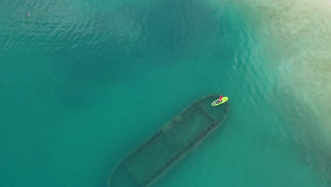 Aerial-view-of-woman-stand-up-paddle-over-rusty-shipwreck-in-clear-turquoise-water-by-tropical-island