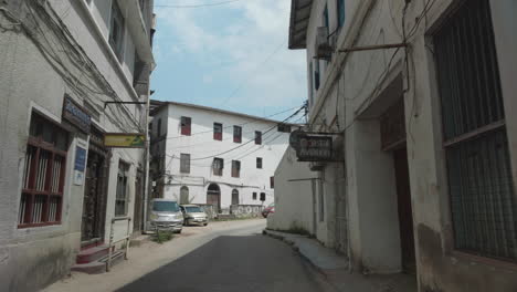 Antigua-Calle-Estrecha-De-La-Ciudad-De-Piedra-De-Zanzíbar-Con-Carteles-De-Bares-Y-Restaurantes-En-Las-Paredes-Y-Coches-Que-Pasan