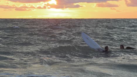 Surfers-on-surfboards-paddling-over-waves-near-the-Baltic-sea-Karosta-beach-at-Liepaja-during-a-beautiful-vibrant-sunset-at-golden-hour,-medium-slow-motion-shot-from-a-distance