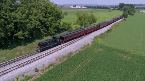 Una-Vista-Aérea-De-Una-Locomotora-Diésel-Tirando-De-Coches-De-Pasajeros-Antiguos-A-Través-De-La-Campiña-Amish