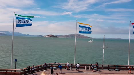 Schuss-Von-Flaggen-Vom-Pier-39,-Die-Bei-Starkem-Wind-Am-Ende-Des-Pier-39-Mit-Der-Insel-Alcatraz-An-Einem-Sommertag-In-San-Francisco,-Kalifornien,-Usa-Wehen