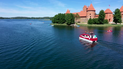 Antena:-Barco-Rojo-Con-Turistas-Navegando-Más-Allá-De-La-Isla-Del-Castillo-De-Trakai-Y-Haciendo-Ondas-En-La-Superficie-Del-Lago