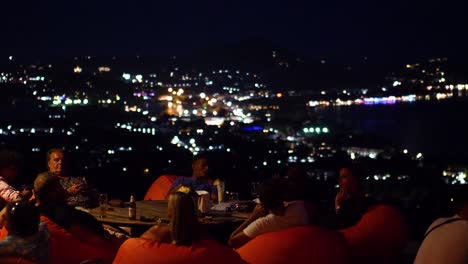 Turistas-Sentados-En-La-Terraza-Infinita-Del-Club-De-La-Selva-En-Samui-Por-La-Noche,-Con-La-Ciudad-De-Chaweng-Al-Fondo