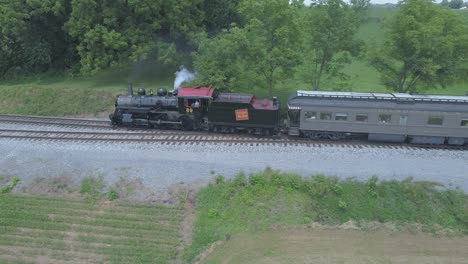 Vista-Aérea-De-Un-Motor-De-Vapor-De-1910-Con-Un-Tren-De-Pasajeros-Que-Echa-Humo-Viajando-A-Lo-Largo-De-La-Campiña-Amish-En-Un-Día-Soleado-De-Verano-Visto-Por-Un-Dron