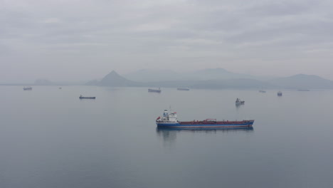 Barcos-En-El-área-De-Espera-De-Anclaje-Con-La-Cresta-De-La-Montaña-En-La-Distancia,-En-Un-Día-Nublado-Y-Tranquilo