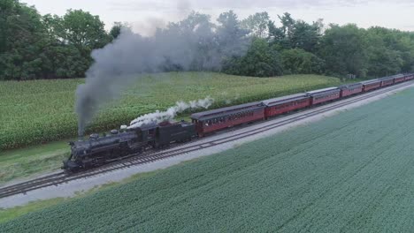 Vista-Aérea-De-Un-Tren-De-Pasajeros-De-Vapor-Que-Sale-De-Una-Estación-De-Tren-En-El-Campo-Amish-En-Un-Día-De-Verano