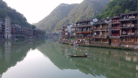 Hombre-Sentado-Y-Remando-En-Un-Viejo-Bote-De-Madera-En-El-Río-Tuo,-Que-Fluye-A-Través-Del-Centro-Del-Casco-Antiguo-De-Fenghuang