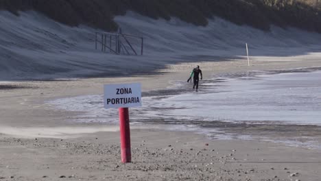 Mann-Im-Neoprenanzug-Mit-Einem-Bodyboard,-Der-Am-Strand-Mit-Der-Beschilderung-Der-Zona-Portuaria-Läuft---Breiter-Slowmo-Schuss