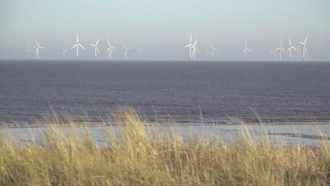 Pastos-Amarillos-De-Playa-En-La-Parte-Superior-De-Las-Dunas-De-Arena-Soplan-En-El-Viento-Mientras-Las-Palas-De-La-Turbina-Giran-En-El-Enorme-Parque-Eólico-Lincs,-Que-Se-Encuentra-A-Ocho-Kilómetros-De-La-Costa-En-El-Mar-Del-Norte-Frente-A-La-Costa-Este-De-Inglaterra